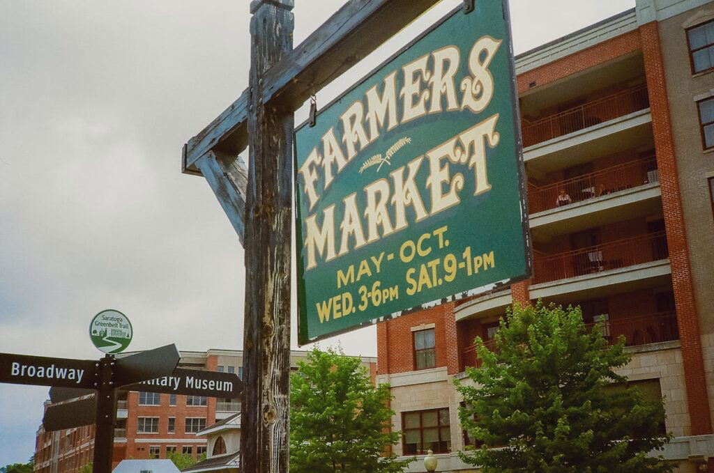Green farmers market sign sits near a Broadway Street sign and brick building in Saratoga Springs, NY