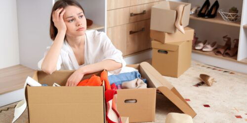A woman sorts through clutter in her home and looks upset while sitting in the floor with boxes of shoes and clothes